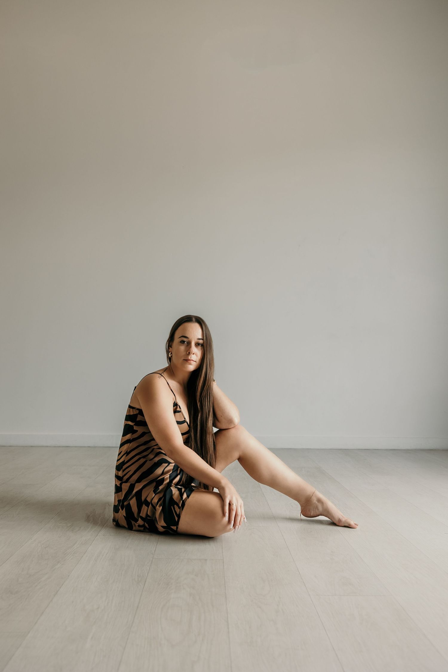Woman in tiger-print dress sitting barefoot on light wood floor against gray wall.