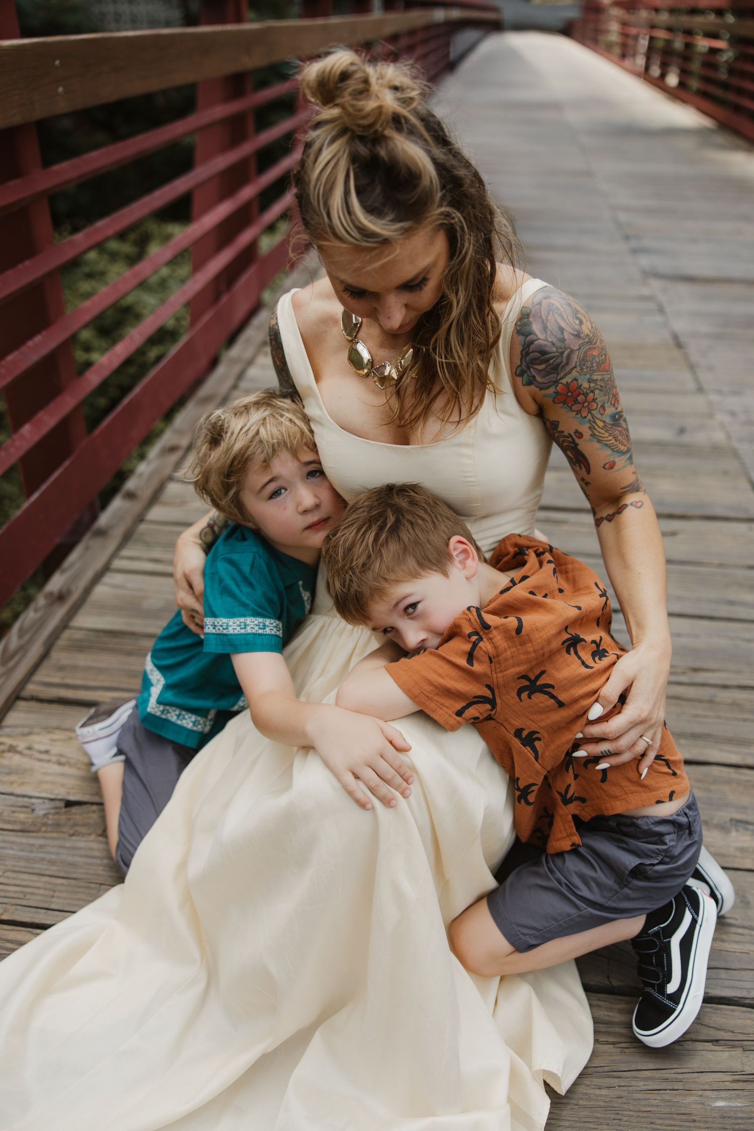 Tattooed mother in white dress embracing two young sons on wooden bridge.