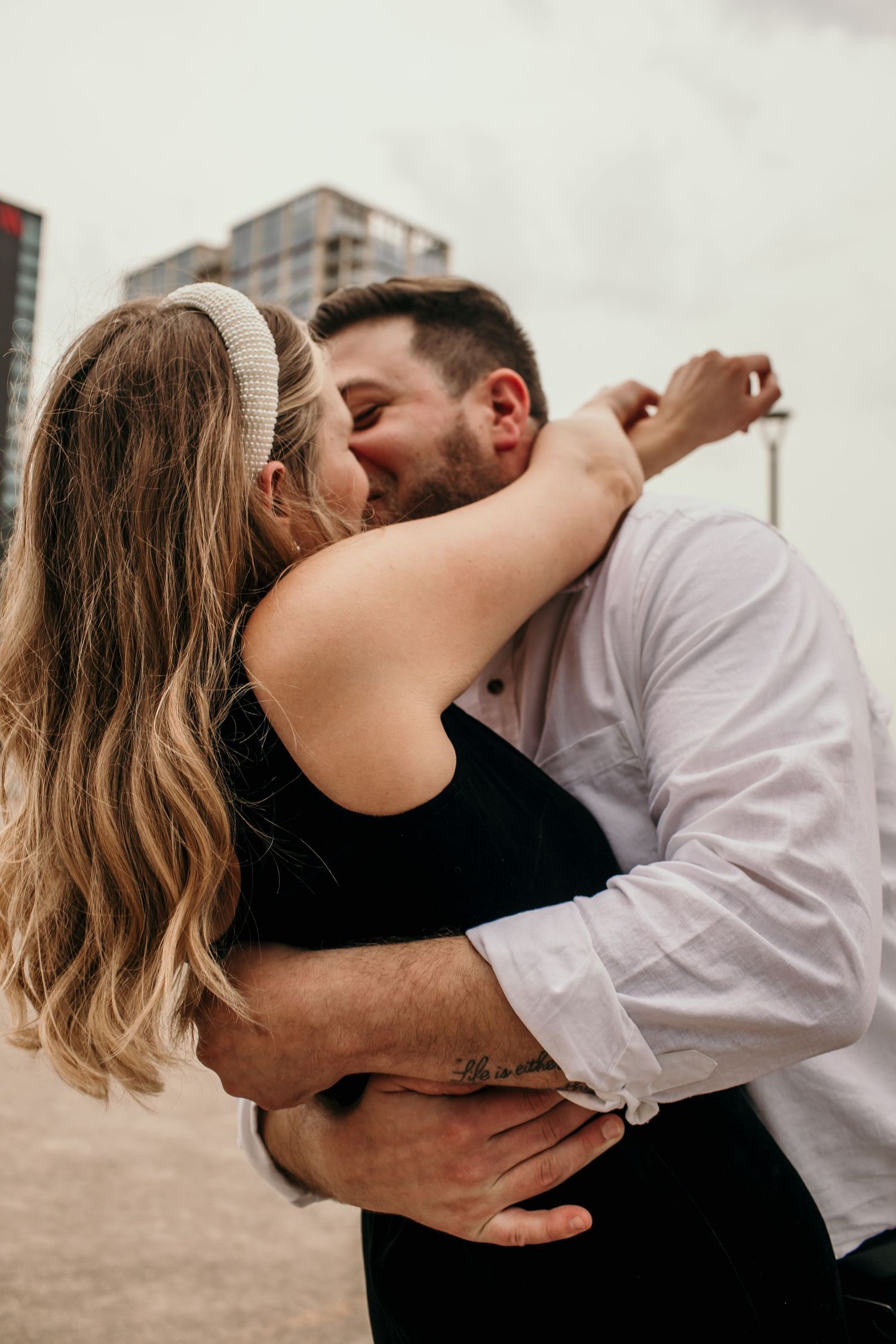 Couple sharing a romantic kiss and embrace outdoors with modern city buildings in the background.