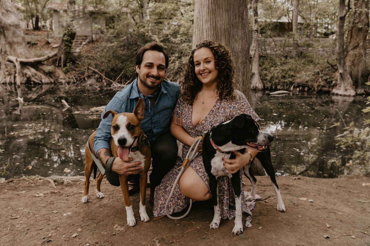 A happy couple crouches with their two dogs in front of a large tree near a woodland creek.