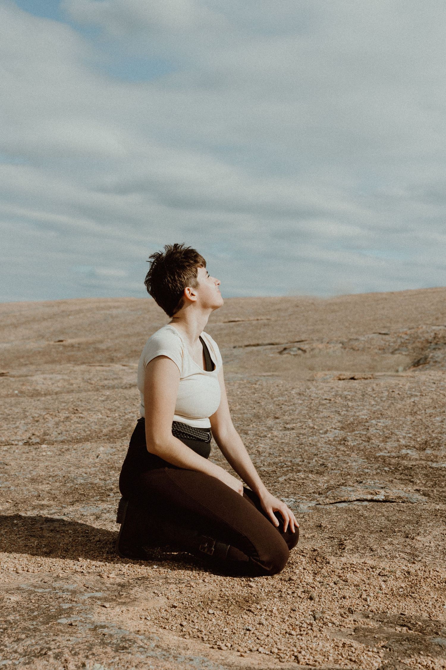 Person kneeling on rocky terrain with eyes closed, facing upward toward cloudy sky in meditative pose.