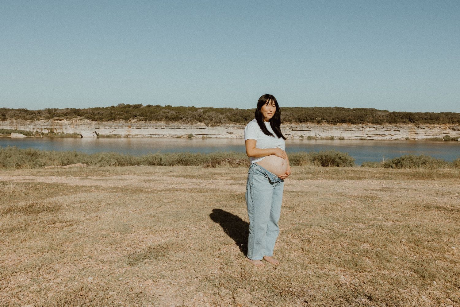 Pregnant woman in white top and jeans standing outdoors near a lake showcasing her baby bump.