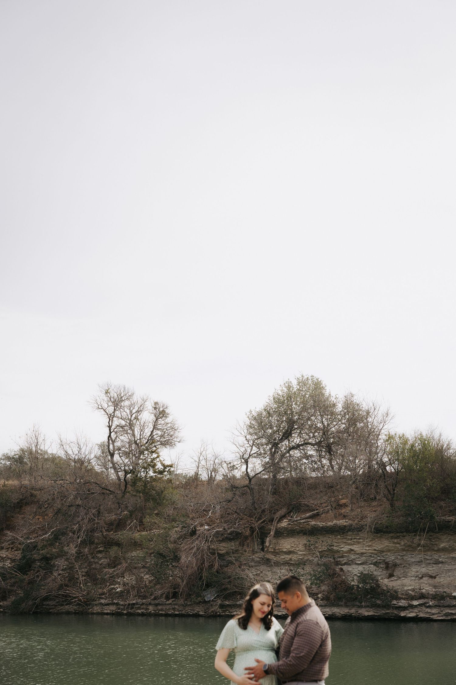 Couple embracing pregnant belly standing near green river with rocky cliffs and bare trees.