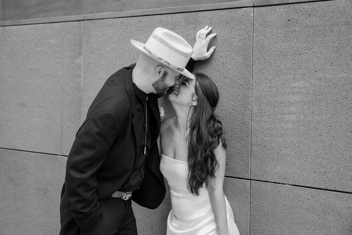 Black and white photo of couple sharing a kiss, bride wearing a wide-brim hat against stone wall.