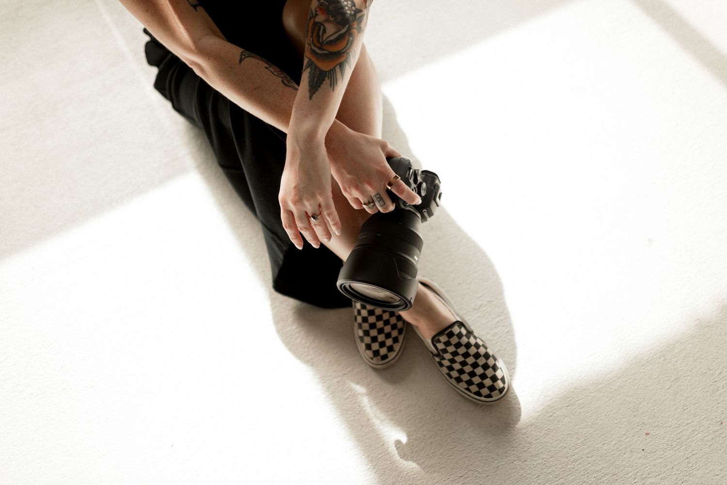 Tattooed hands adjust a checkered Vans slip-on shoe while sitting on a bright white floor.