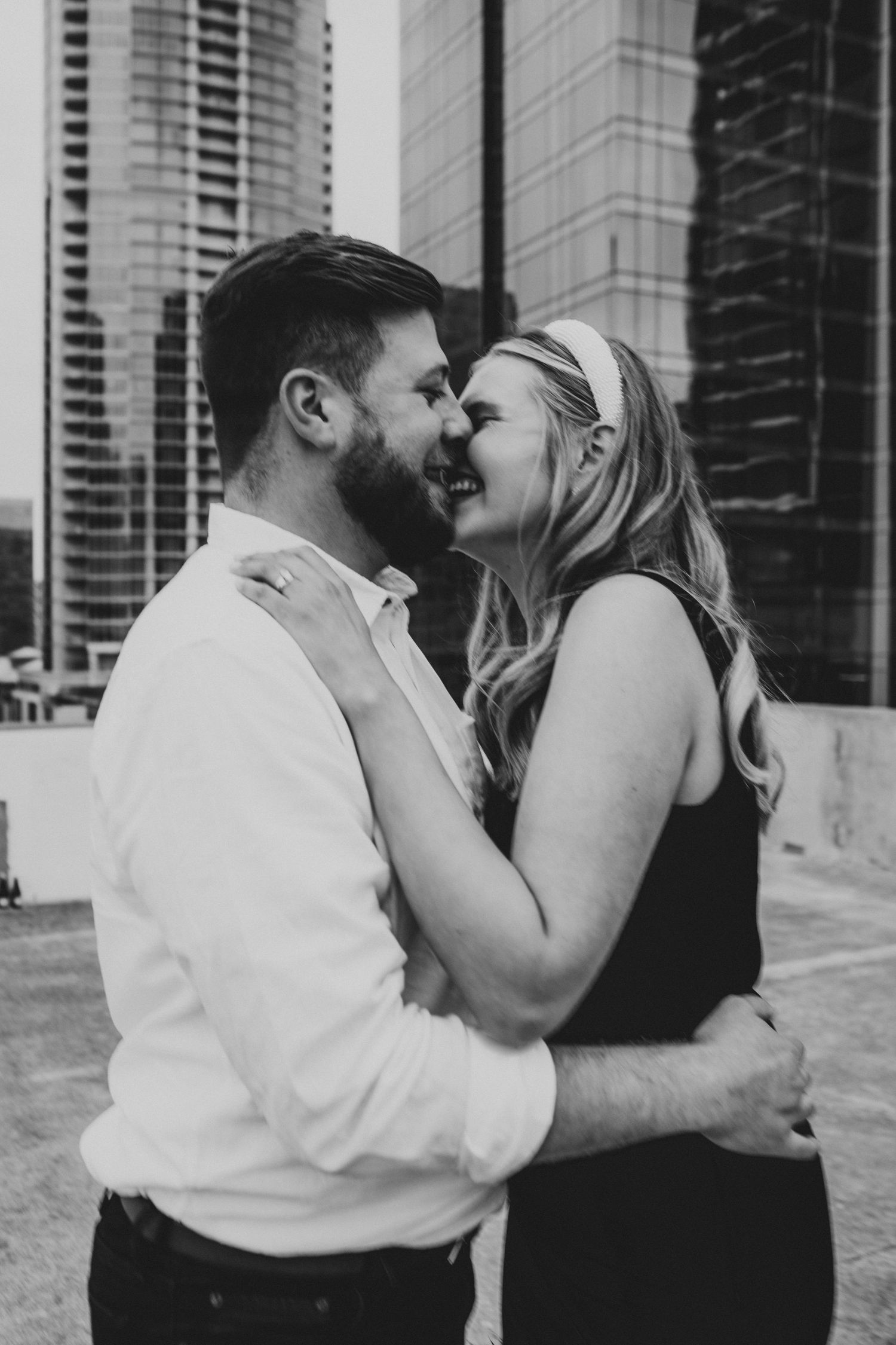 Black and white urban engagement photo of smiling couple embracing with city skyscrapers in background.
