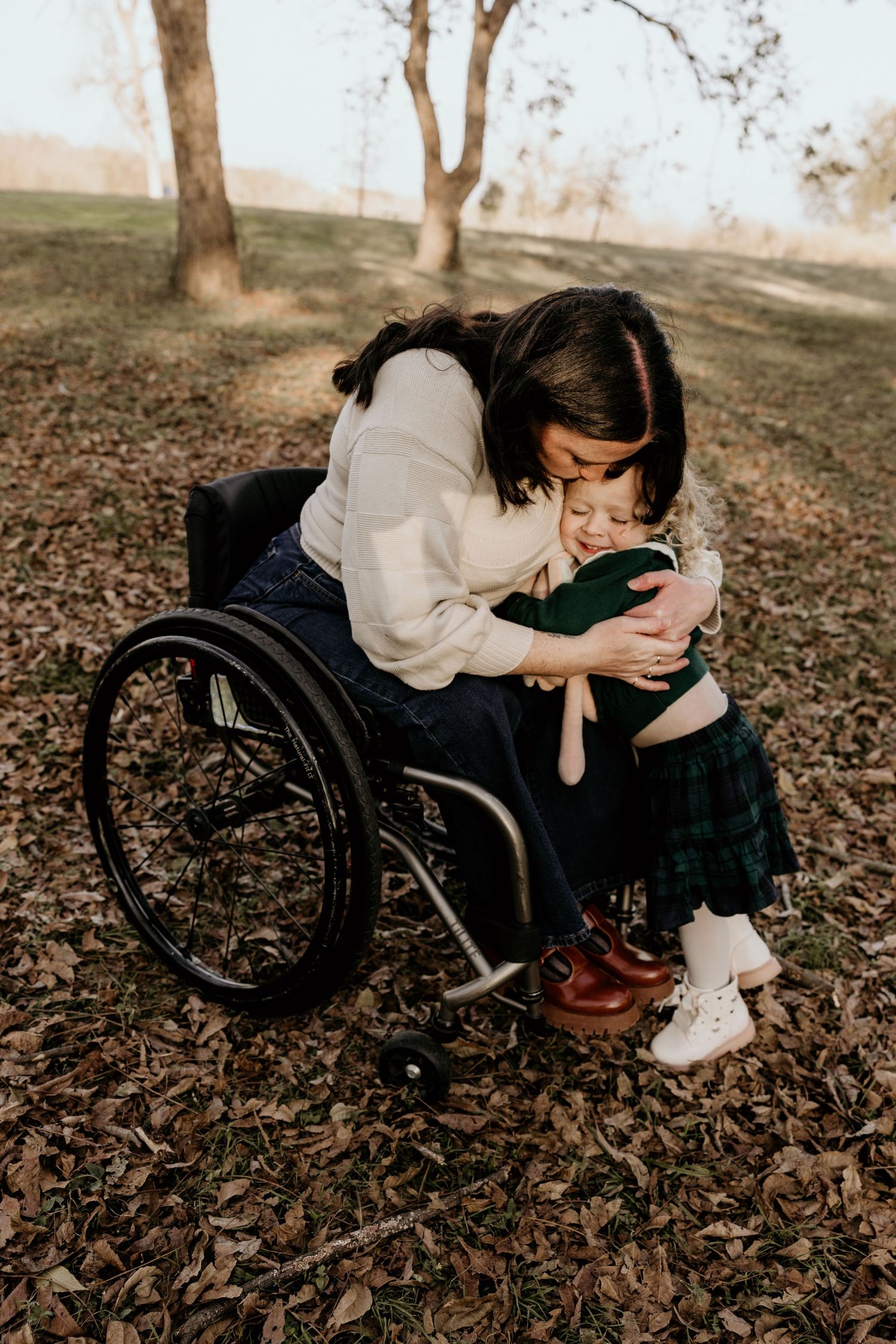 Mother in wheelchair tenderly hugging young curly-haired toddler outdoors among fallen autumn leaves.