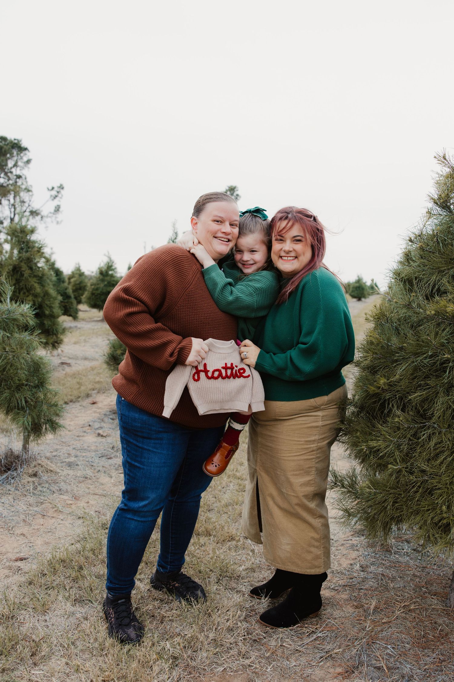 Two moms smiling with daughter holding personalized Hattie bag at Christmas tree farm.