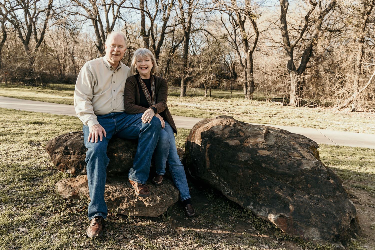 Happy older couple sitting together on large rocks in a park with trees in background.