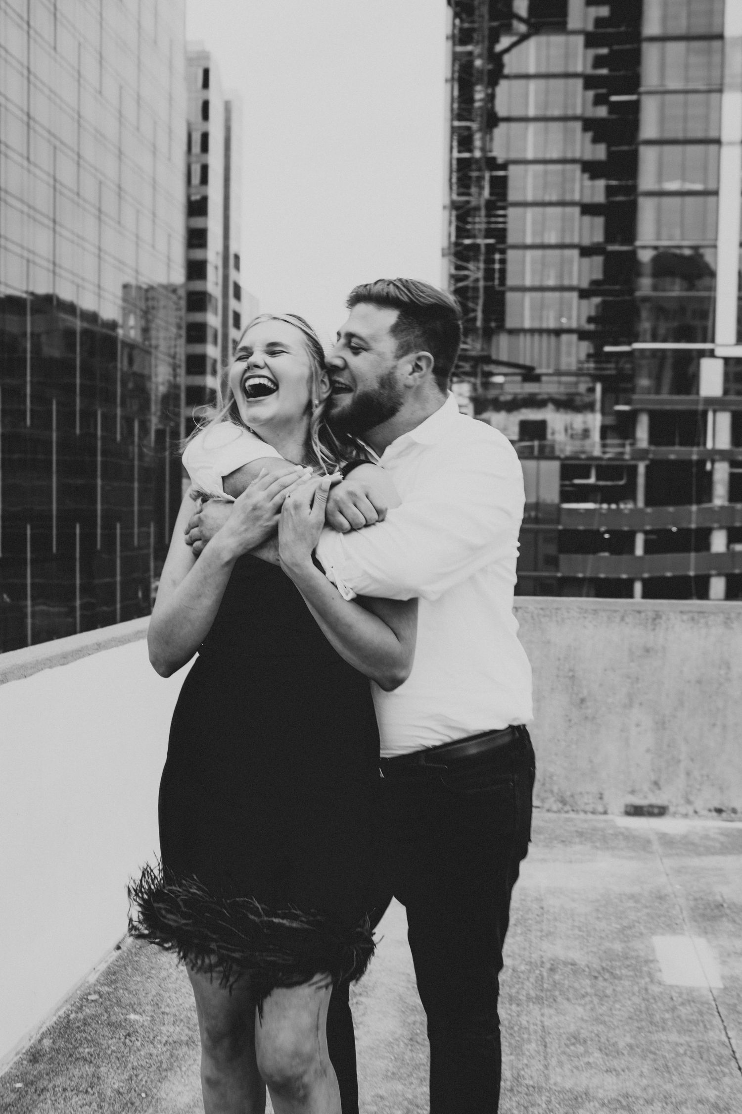 Black and white photo of laughing couple embracing on urban rooftop with city buildings behind.