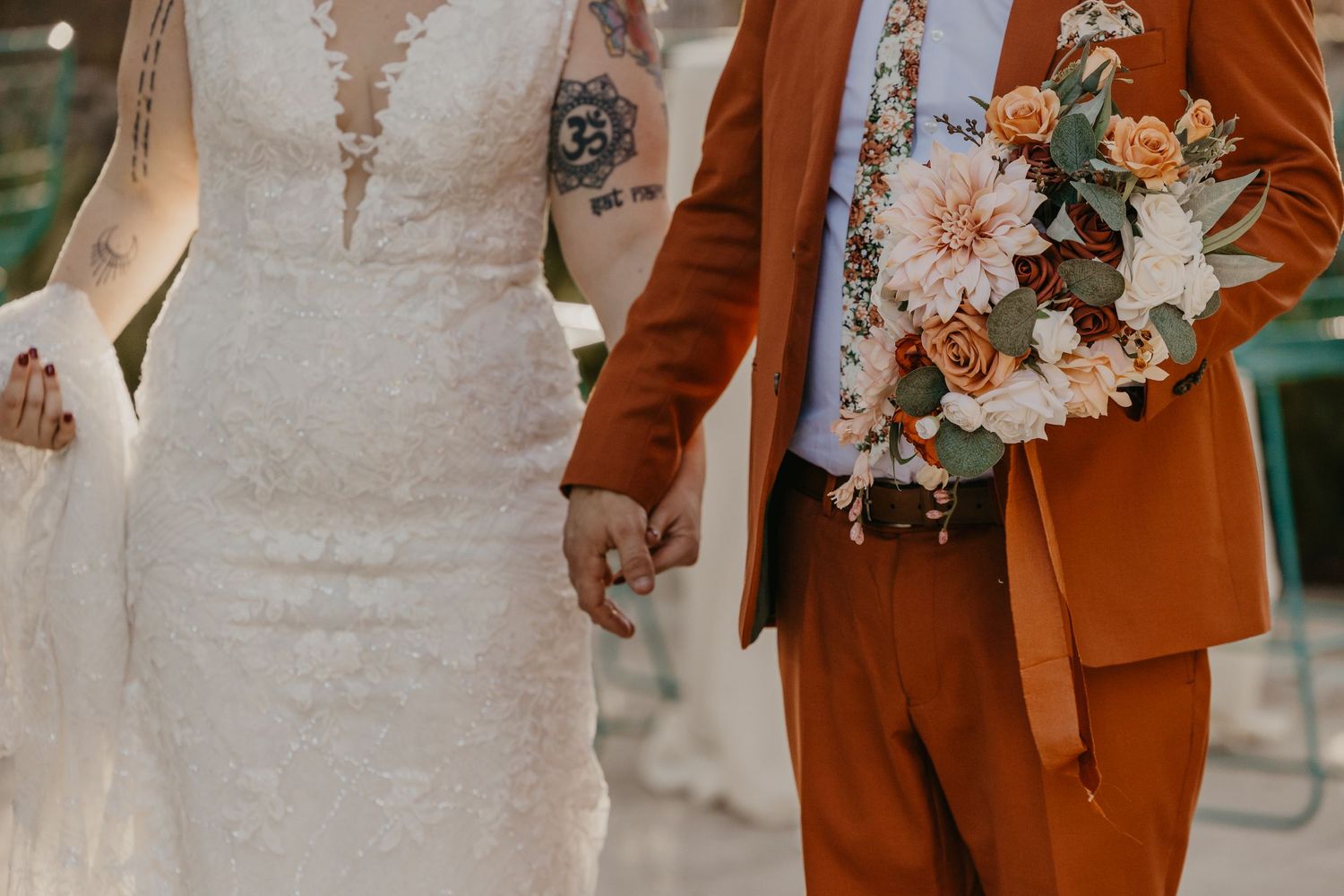 Close-up of bride and groom holding hands, groom in burnt orange suit holding a peach and ivory floral bouquet.