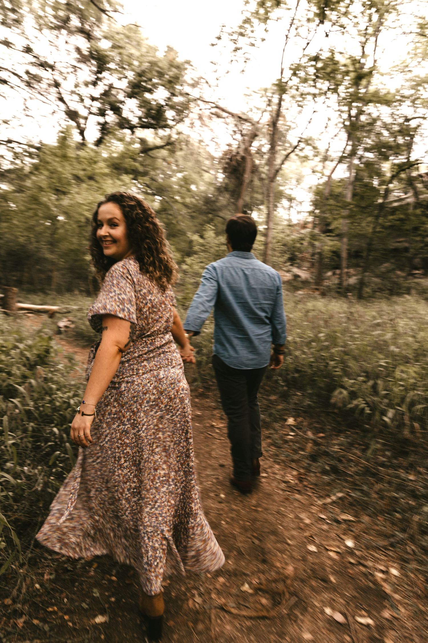 Woman in floral maxi dress smiles back at camera while holding hands with man on forest path.