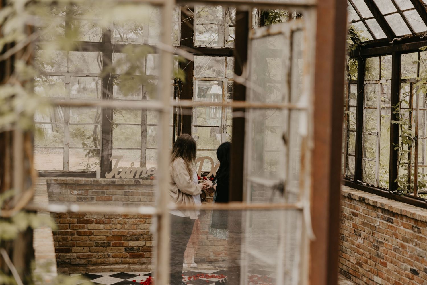 Two women sharing a tender moment inside a vintage greenhouse with brick walls.