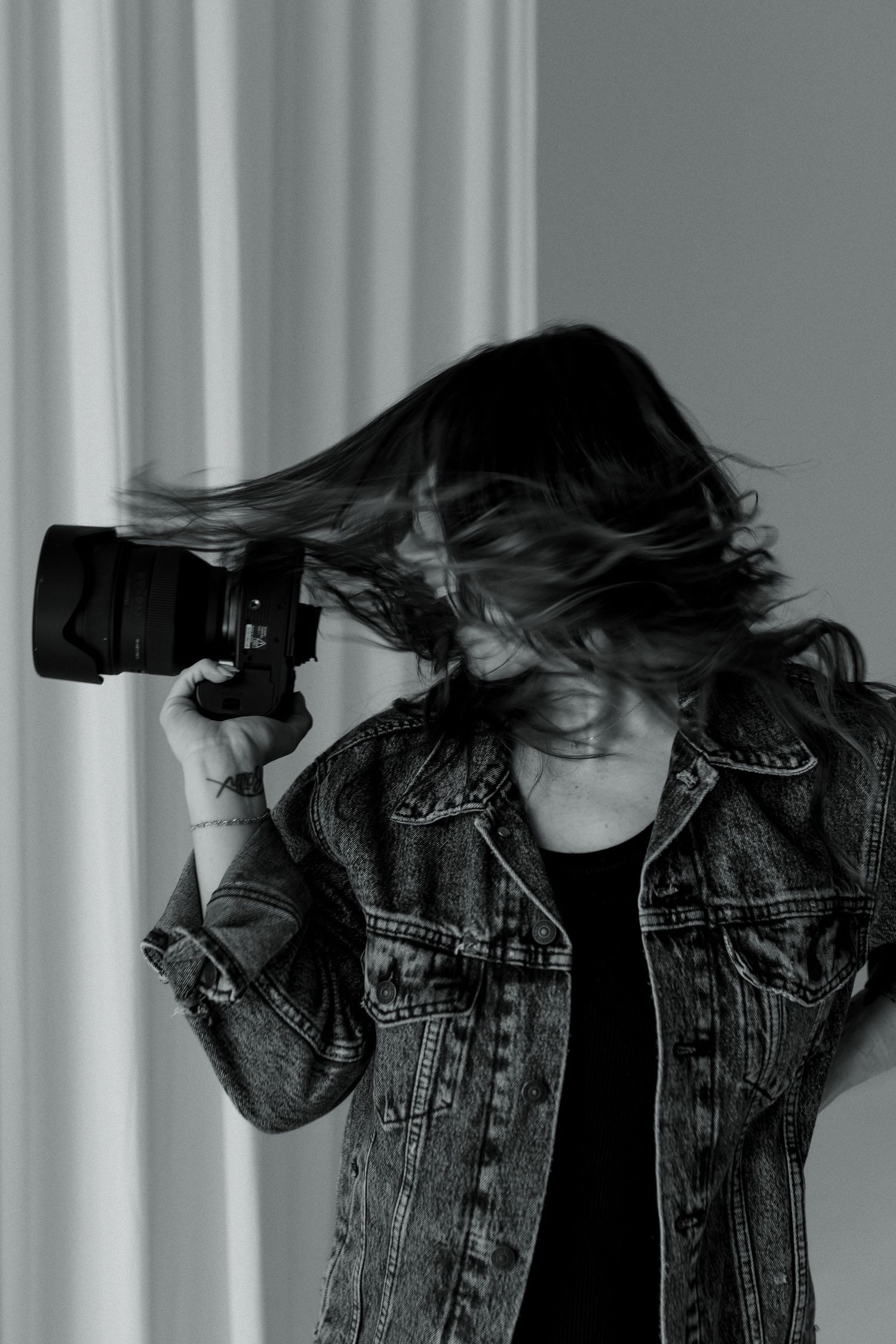 Black and white photo of a woman in a denim jacket holding a camera, her hair dramatically blowing across her face.