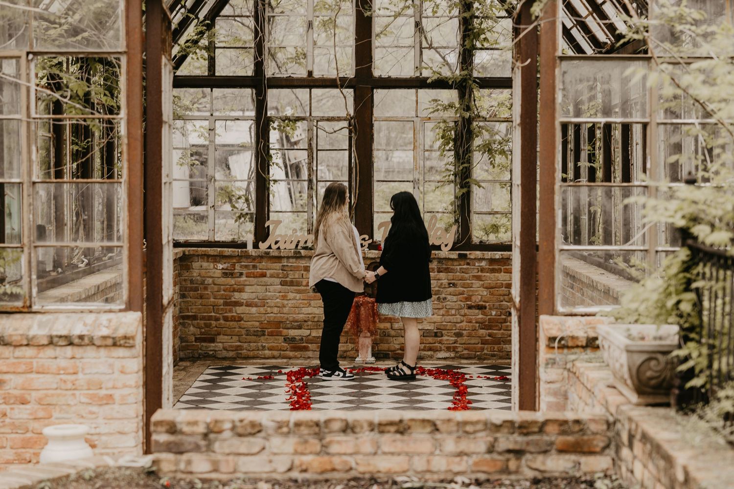 Two women standing together inside a rustic glass greenhouse with a checkered floor.