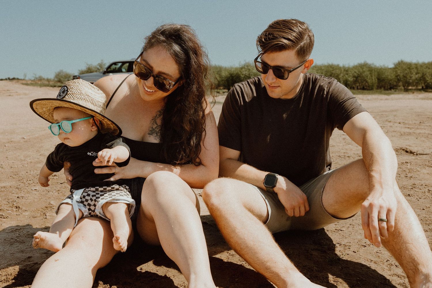 Family with sunglasses relaxing on sandy beach with their baby wearing a straw hat on a sunny day.