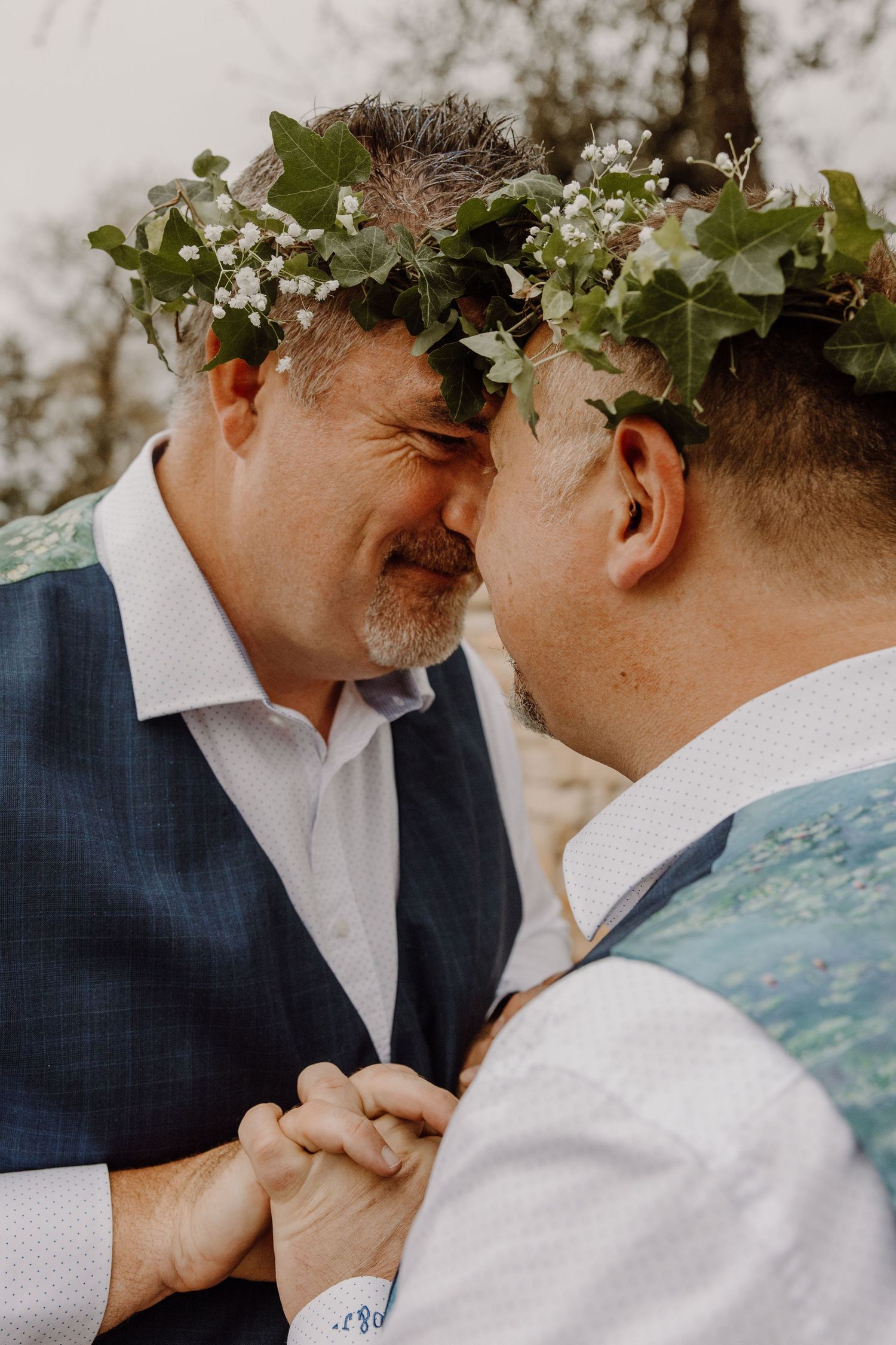 Two grooms wearing floral crowns touching foreheads and holding hands in an intimate outdoor moment.