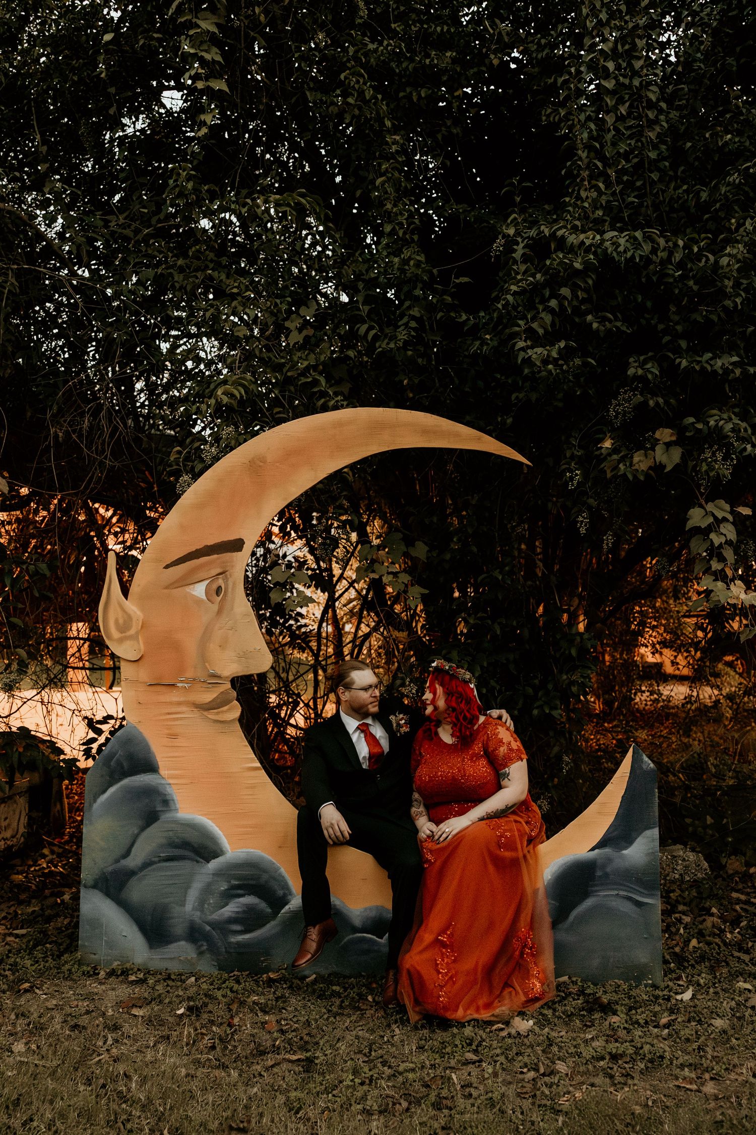 Wedding couple poses beside decorative crescent moon prop surrounded by lush green trees at dusk.