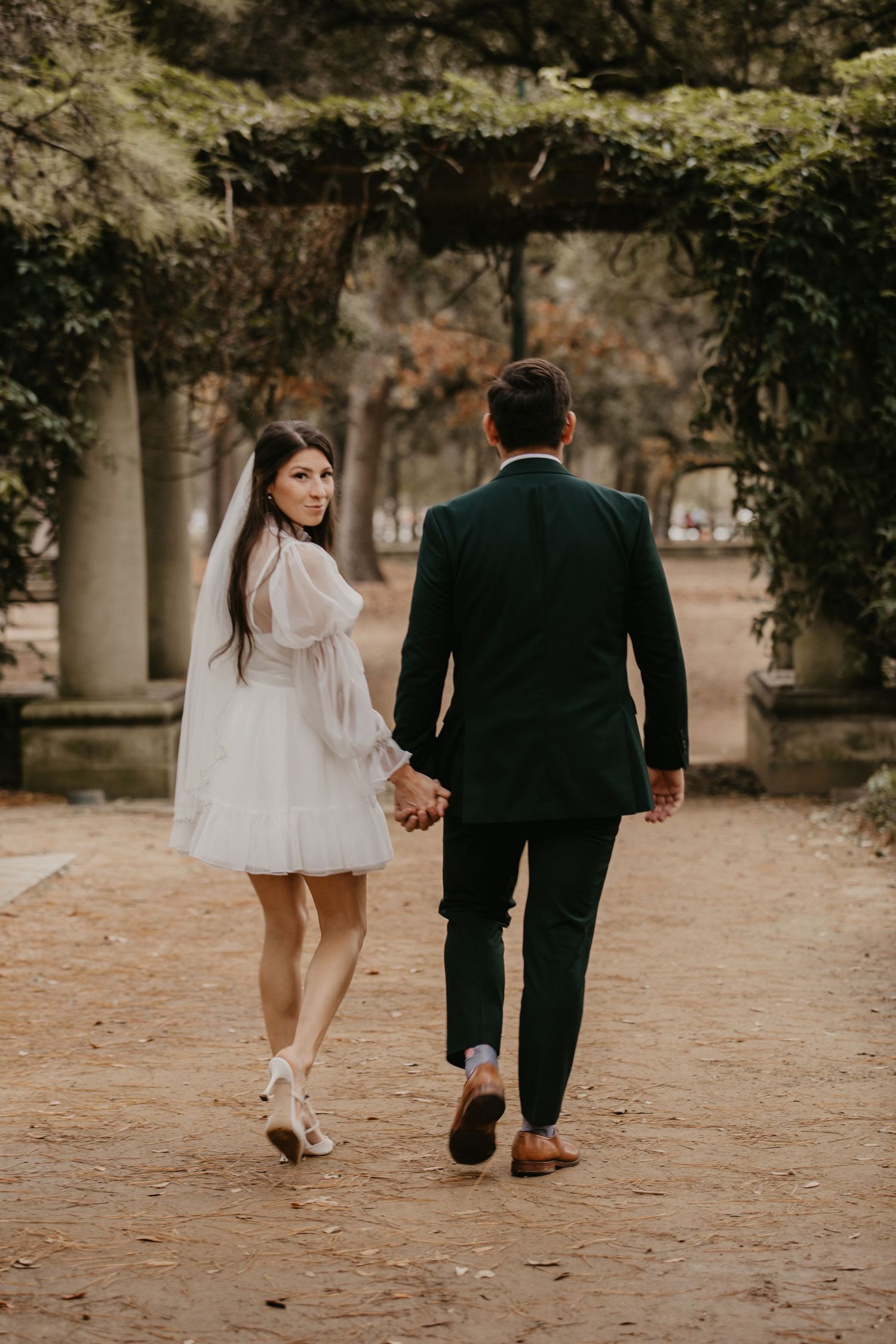 Bride in short white dress and veil holds grooms hand walking away on a tree-lined garden path.