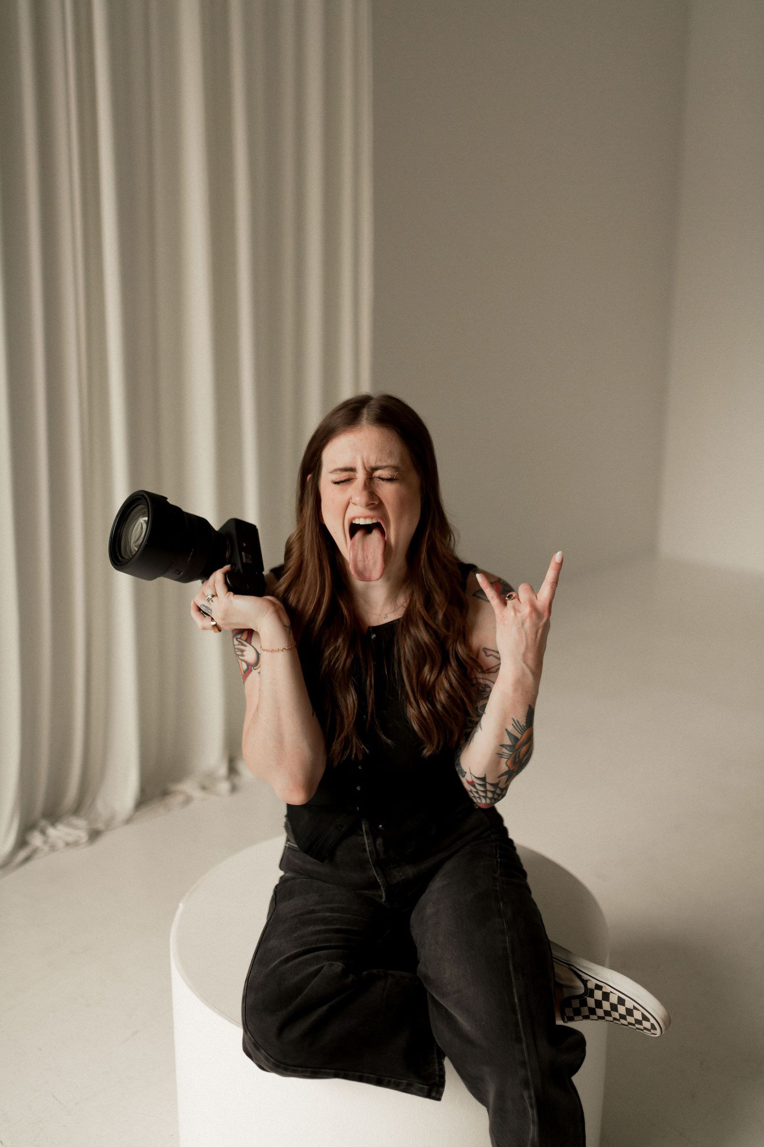 Woman sitting on white pedestal holding camera making rock sign with tongue out in bright studio setting.