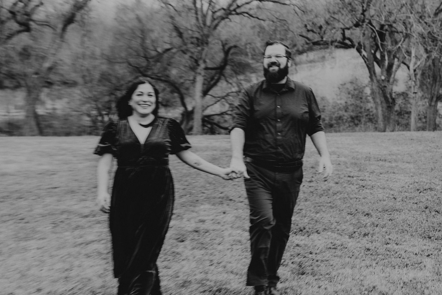 Black and white photo of a couple holding hands and walking through a grassy field with bare trees.