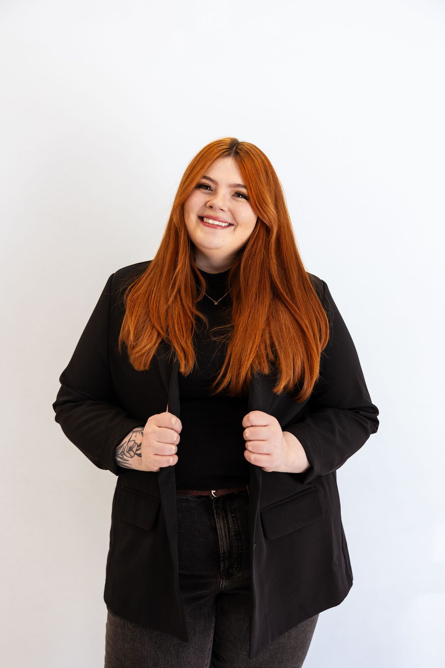 Smiling redheaded woman with tattoos wearing black blazer posing against white background.