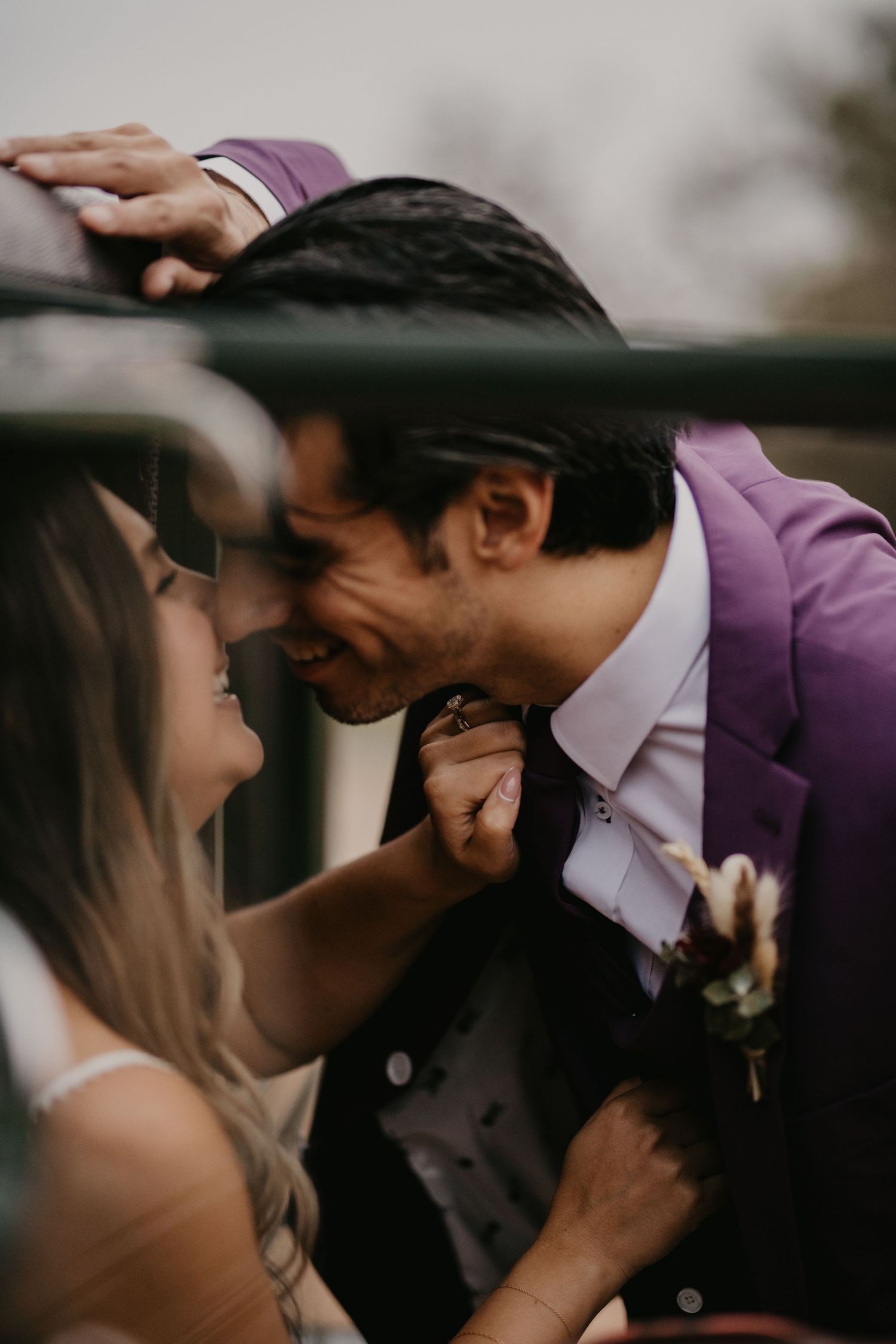 A bride and groom in a purple suit share an intimate forehead touch moment through a blurred foreground.