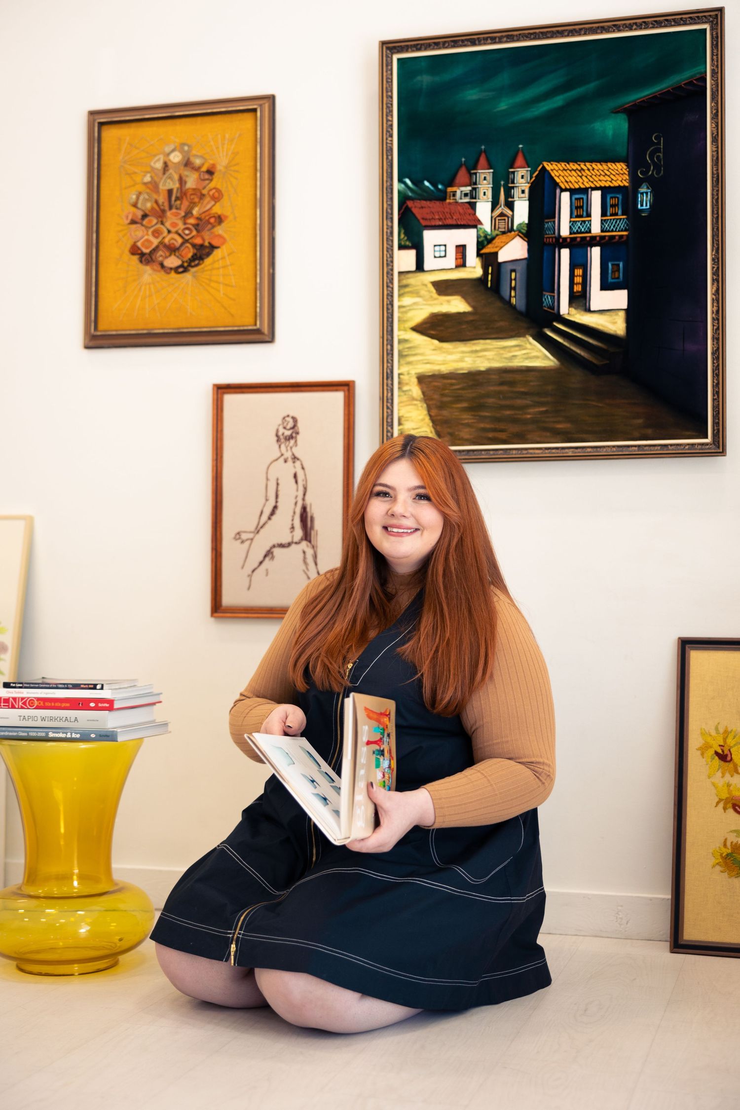 Smiling redheaded woman sitting cross-legged on floor holding artwork among vintage painting collection.