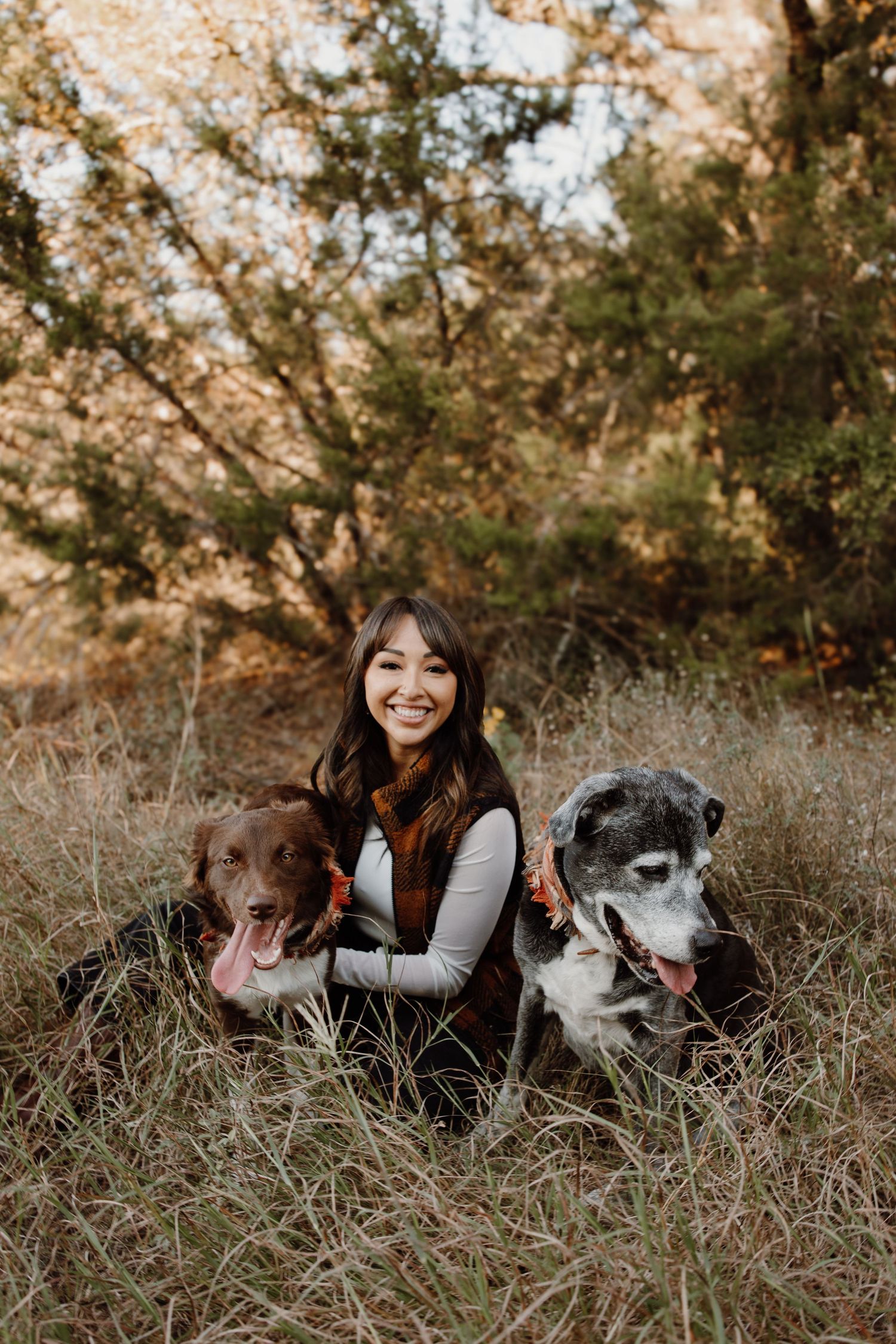 Smiling woman posing with two dogs sitting in tall grass surrounded by autumn foliage.