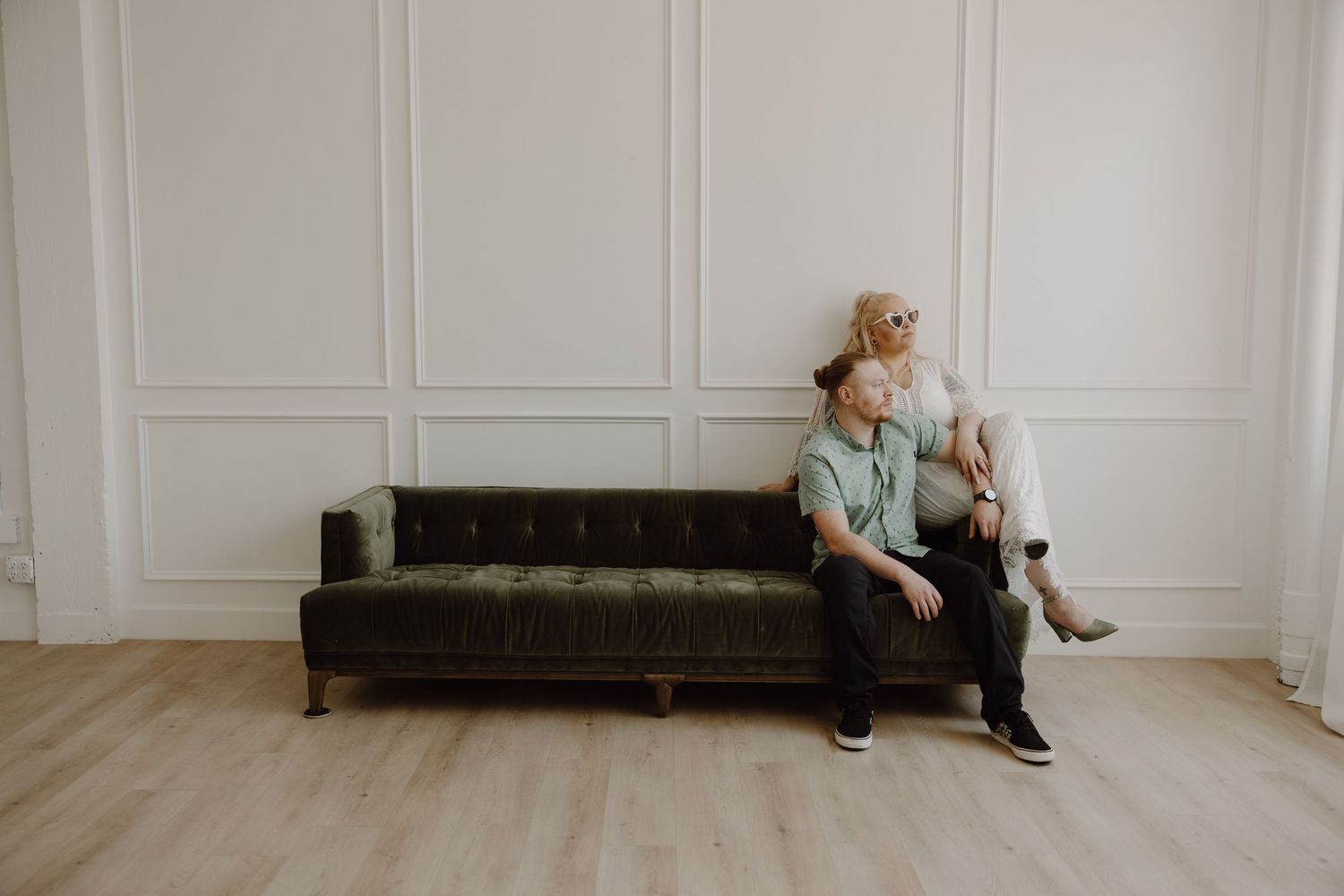 Couple sitting together on green velvet sofa against white paneled wall in bright studio.