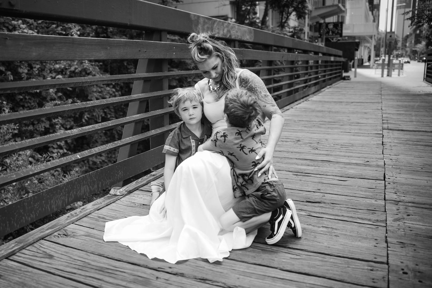 Black and white portrait of a mother embracing her two children on a wooden boardwalk bridge.