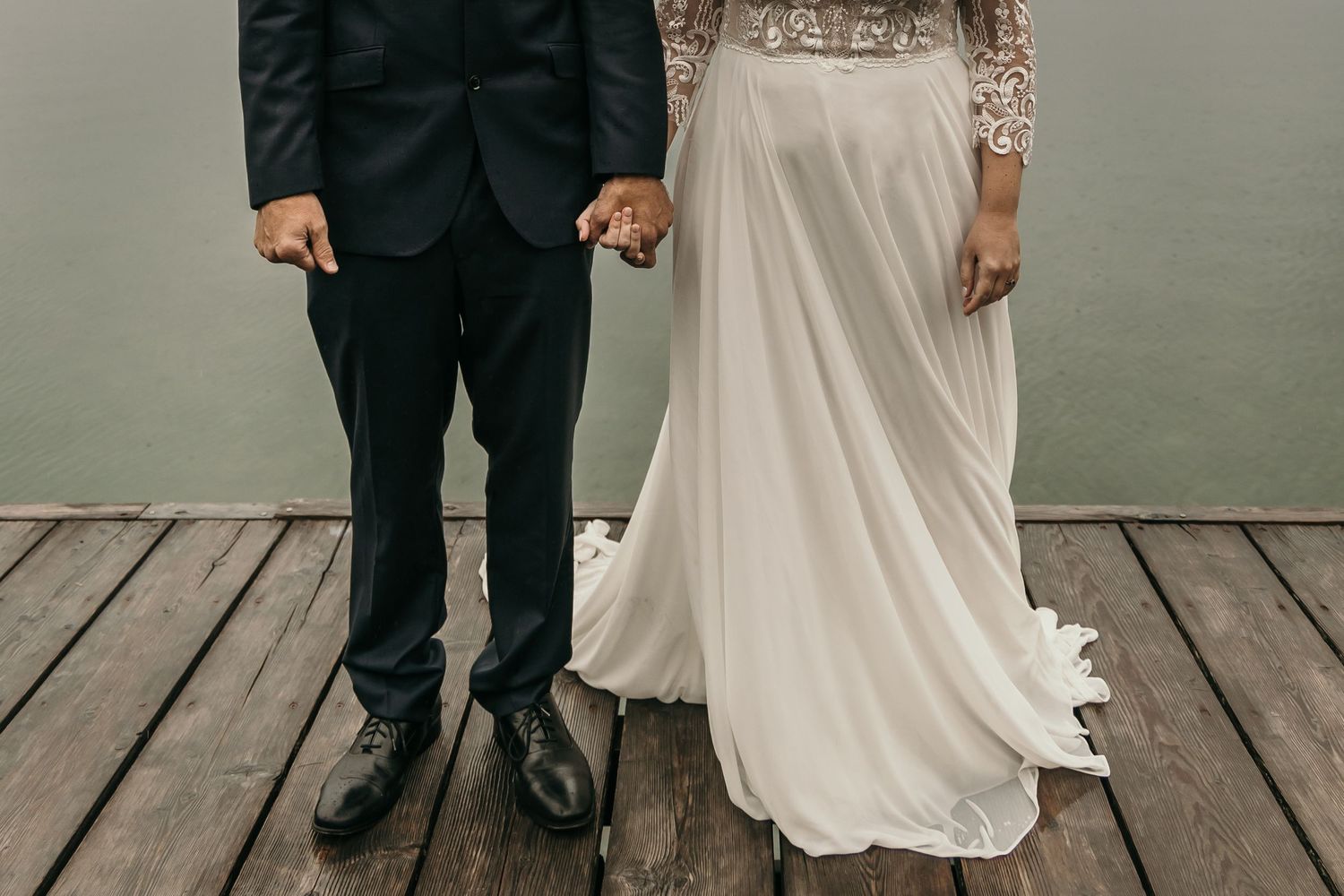 Close-up of bride in flowing white gown and groom in black suit holding hands on a wooden dock.