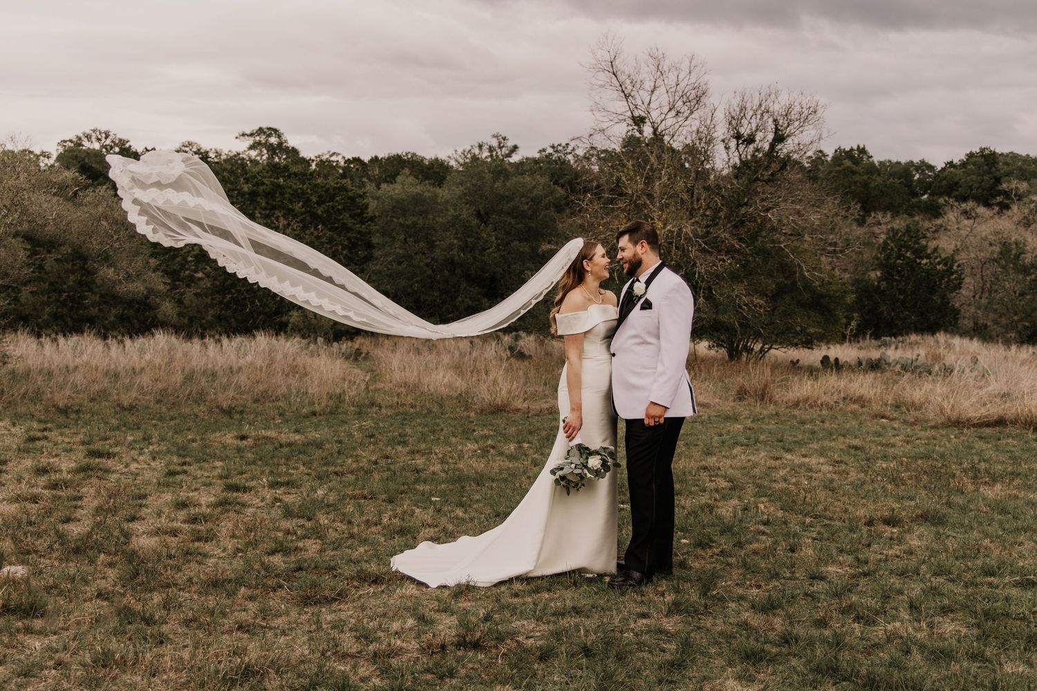 A couple shares a romantic embrace in a field as the bride's veil dramatically billows in the wind.