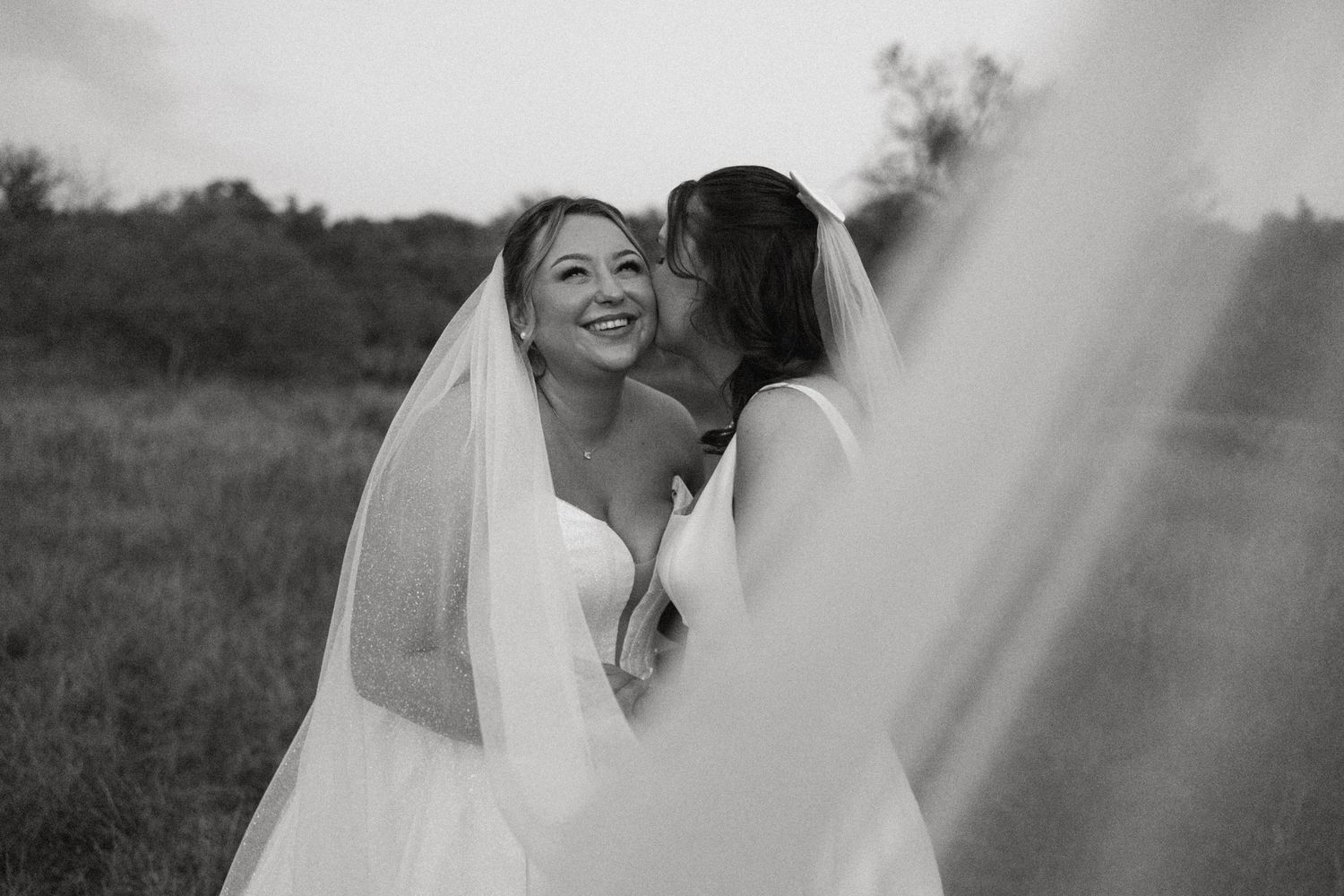 Two brides share a joyful kiss and embrace outdoors in a romantic black and white wedding photo.