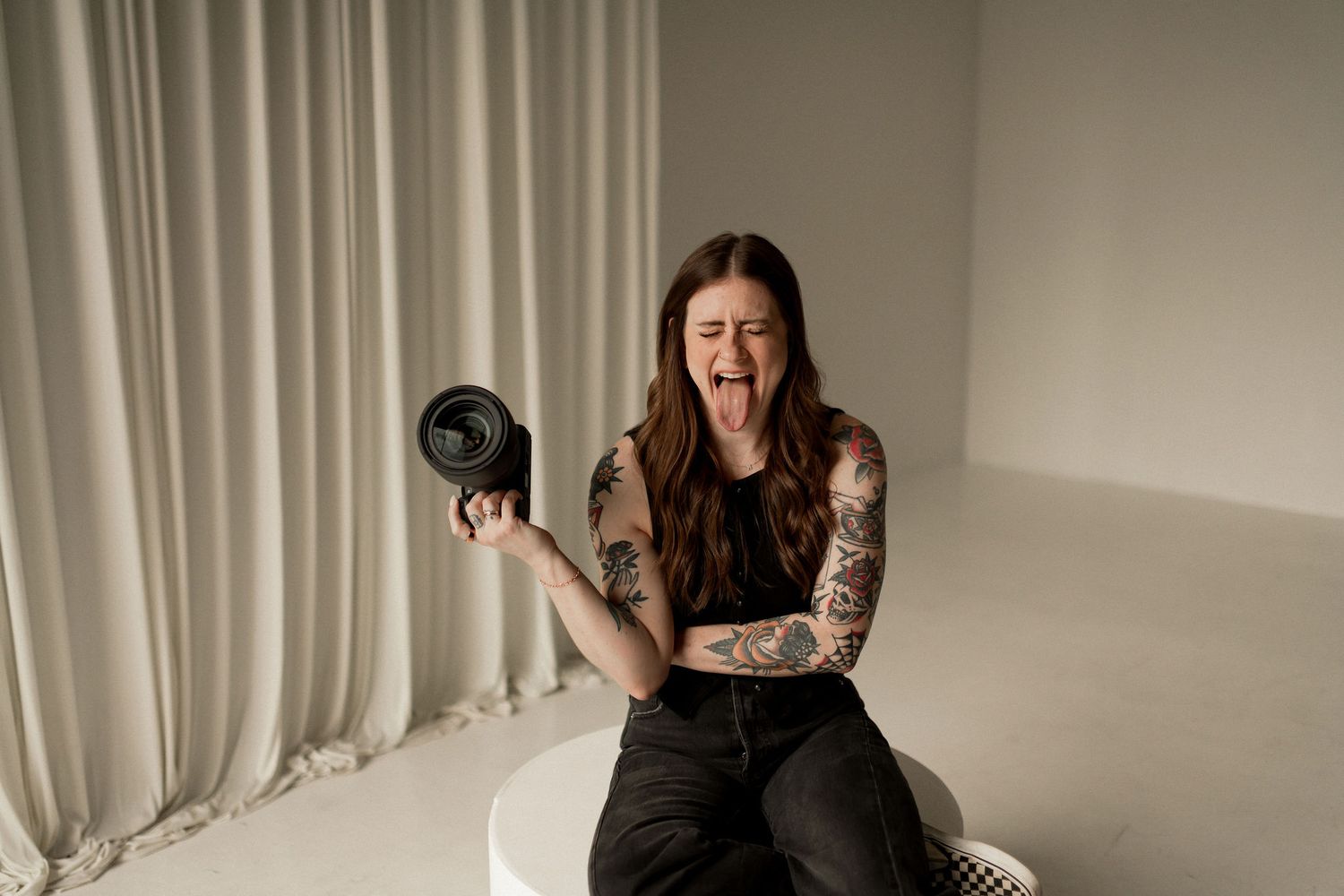 Tattooed woman sitting on white stool, holding a camera, mouth open screaming, wearing black outfit against white curtain.