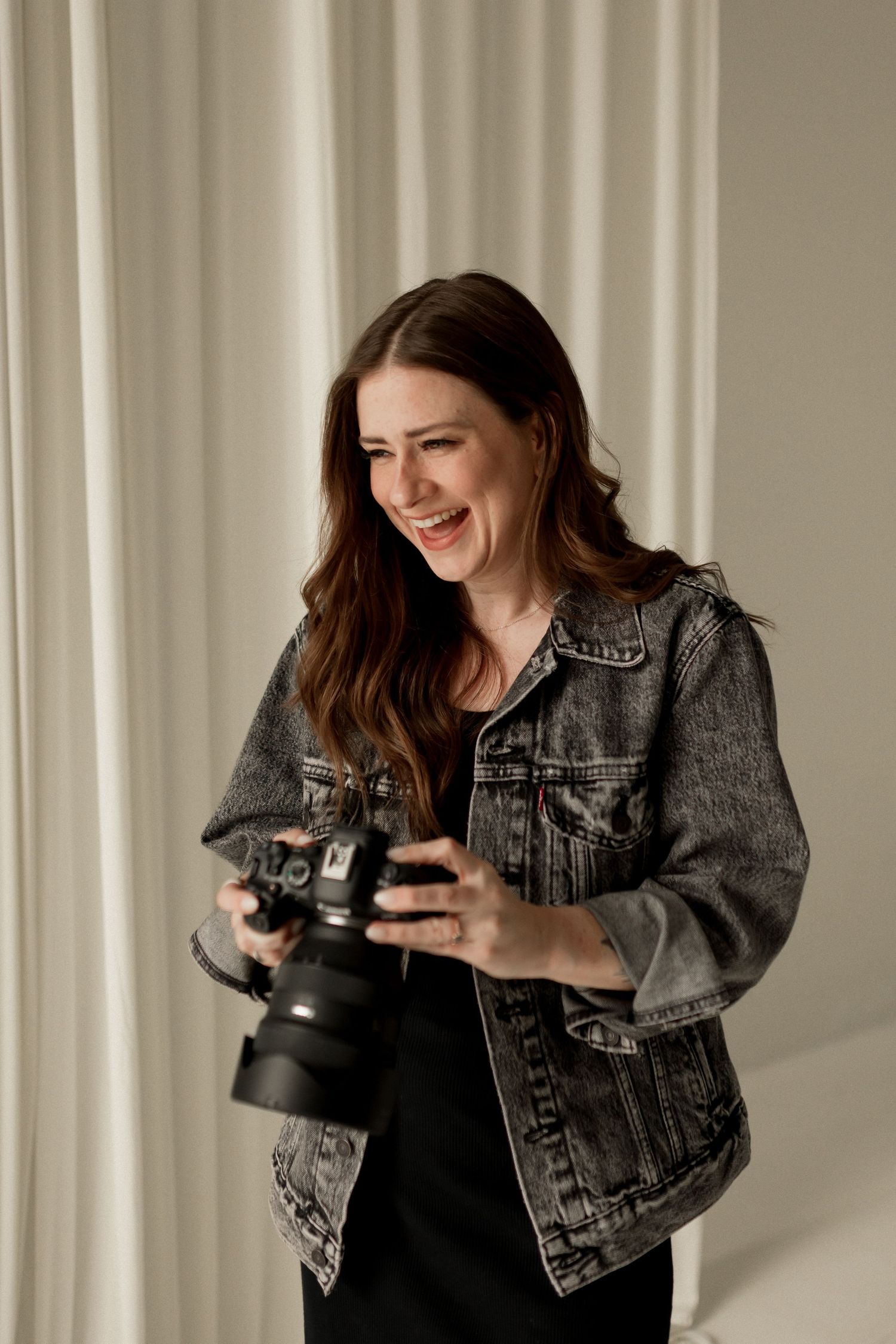Happy female photographer in denim jacket reviewing photos on camera in bright studio.