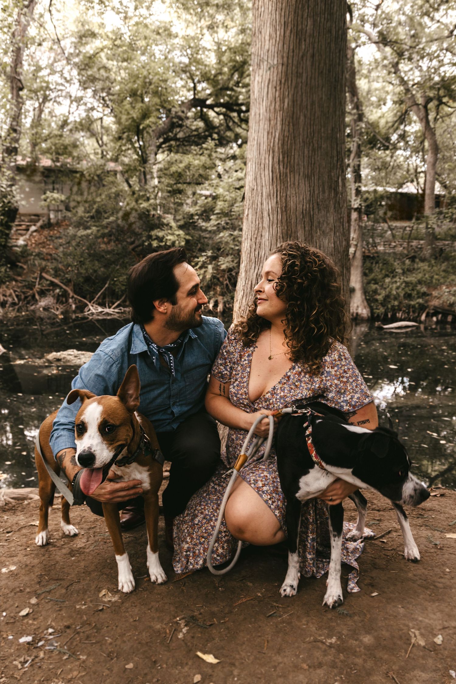 A smiling couple sits with two dogs near a tree by a creek in a lush wooded setting.