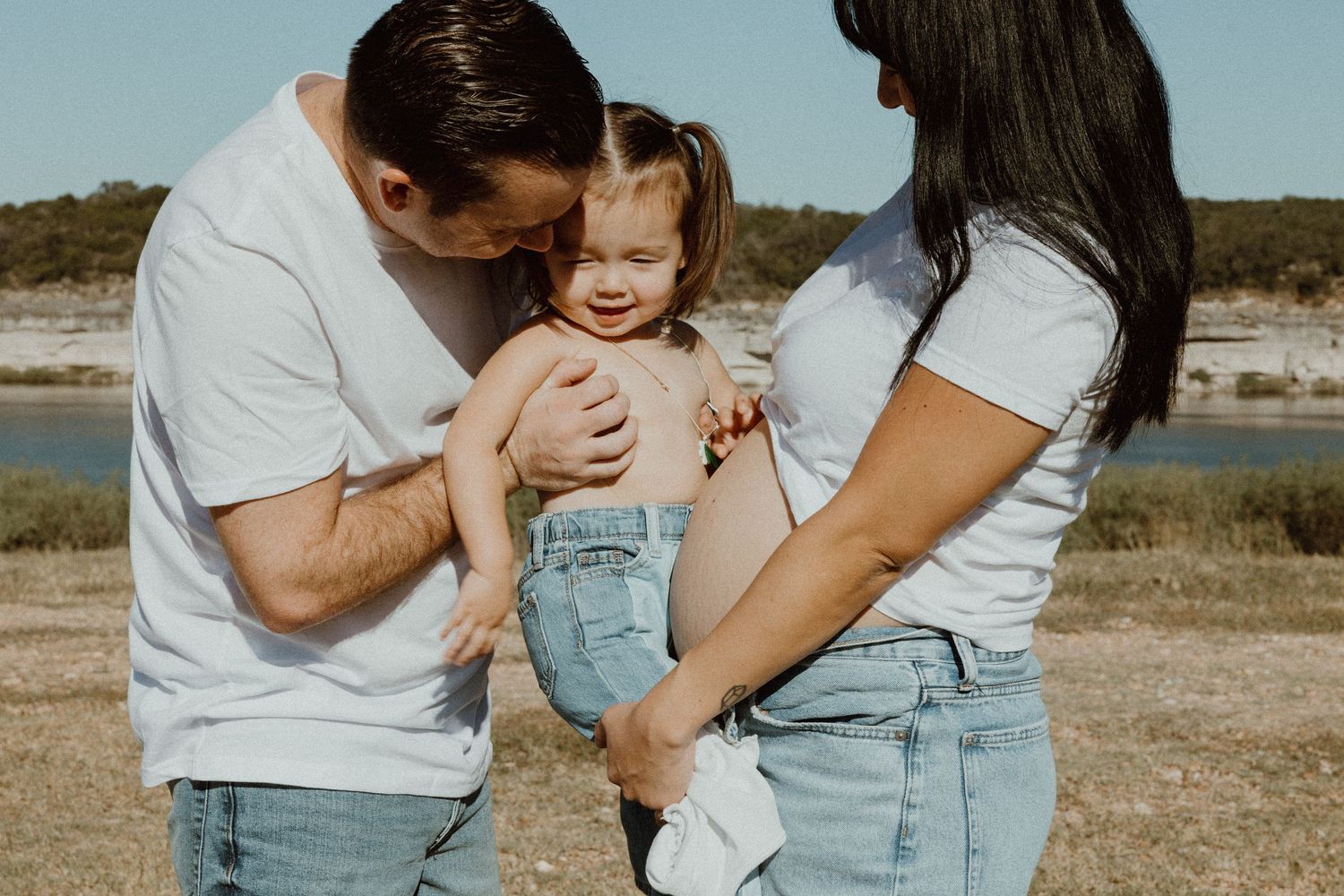 Parents in matching white tees and jeans lovingly holding their smiling toddler near a lake.