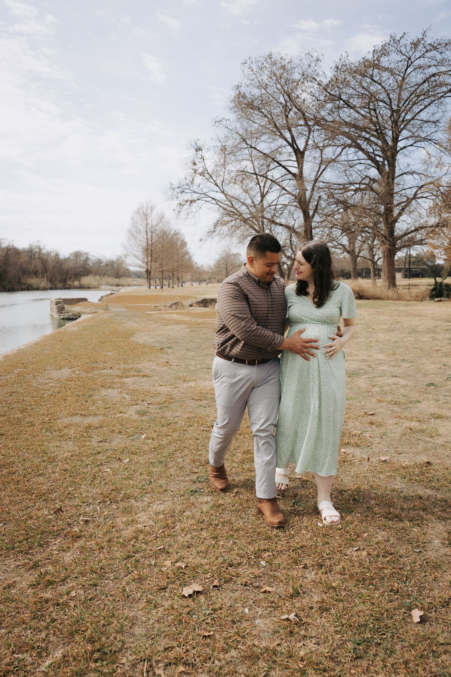 Pregnant couple in mint green dress and plaid shirt walking near river in autumn park.