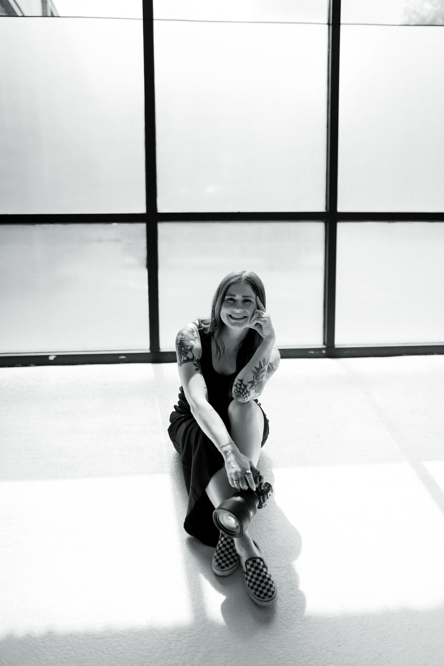 Black and white photo of smiling tattooed woman sitting on bright studio floor with camera.