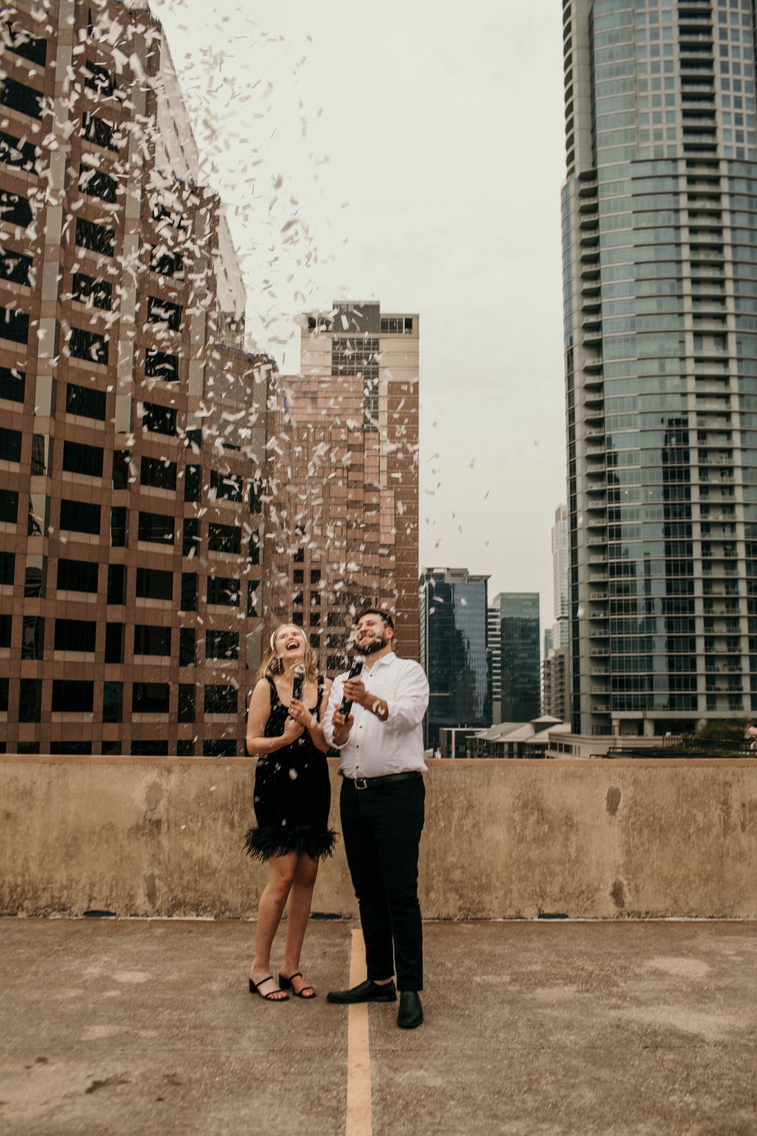 Excited couple celebrating with confetti explosion on urban rooftop surrounded by city skyscrapers.
