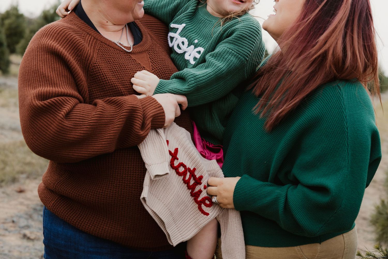 Close-up of two mothers holding child and personalized baby sweater at Christmas tree farm.