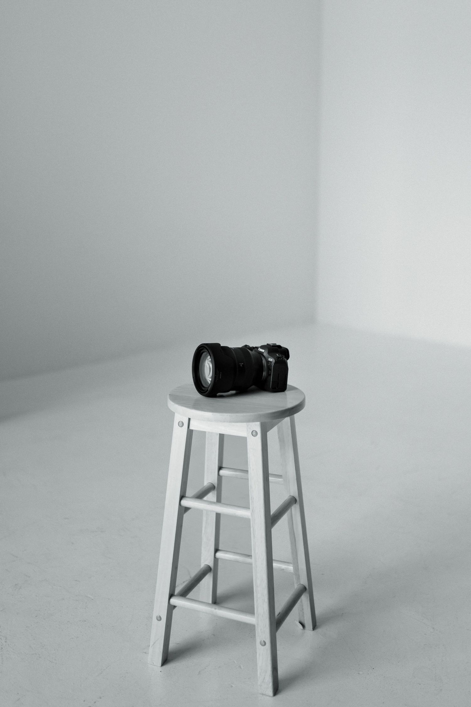 Black DSLR camera with zoom lens resting on a white wooden stool in a minimalist white photography studio.