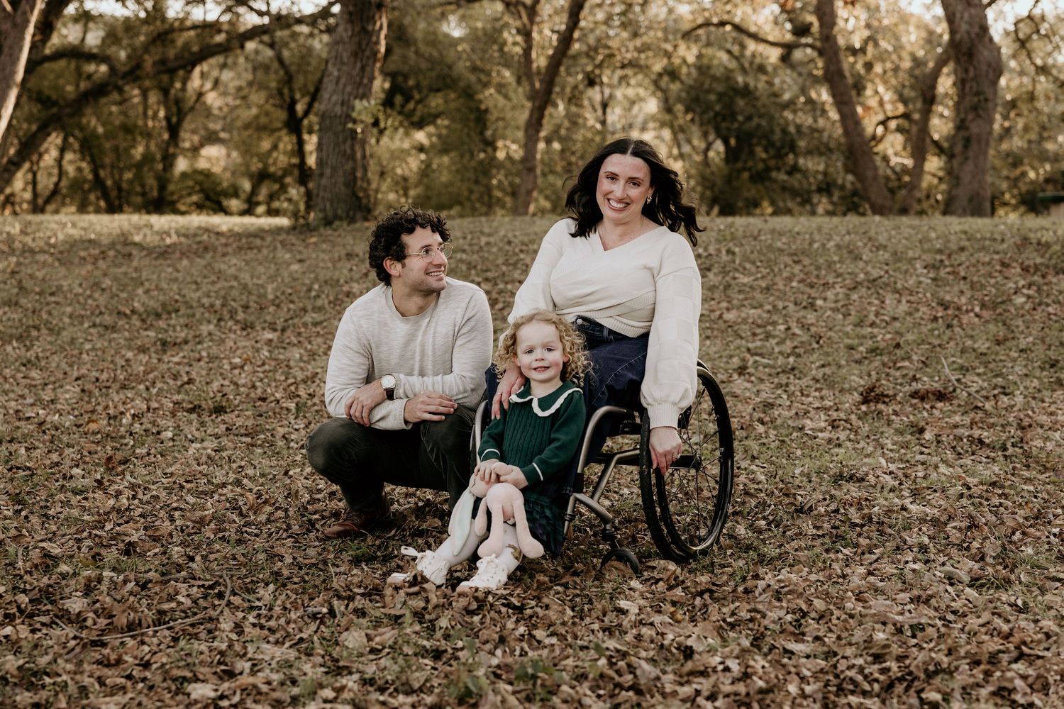Smiling family with mother in wheelchair, father, and young daughter posing together on fallen leaves in a park.