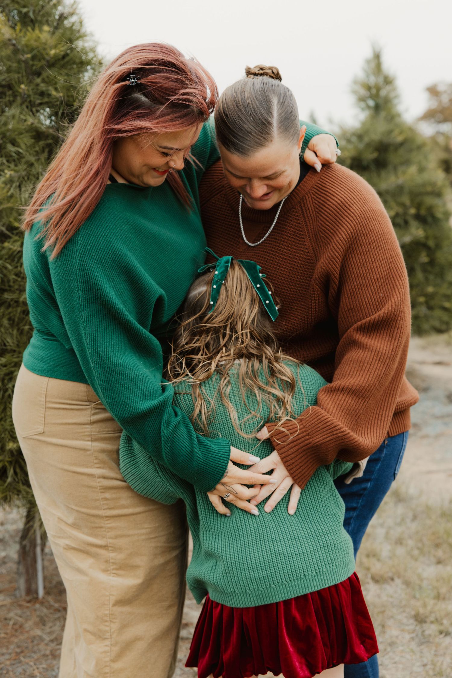 Two women in green and brown sweaters hugging young girl from behind at Christmas tree farm.