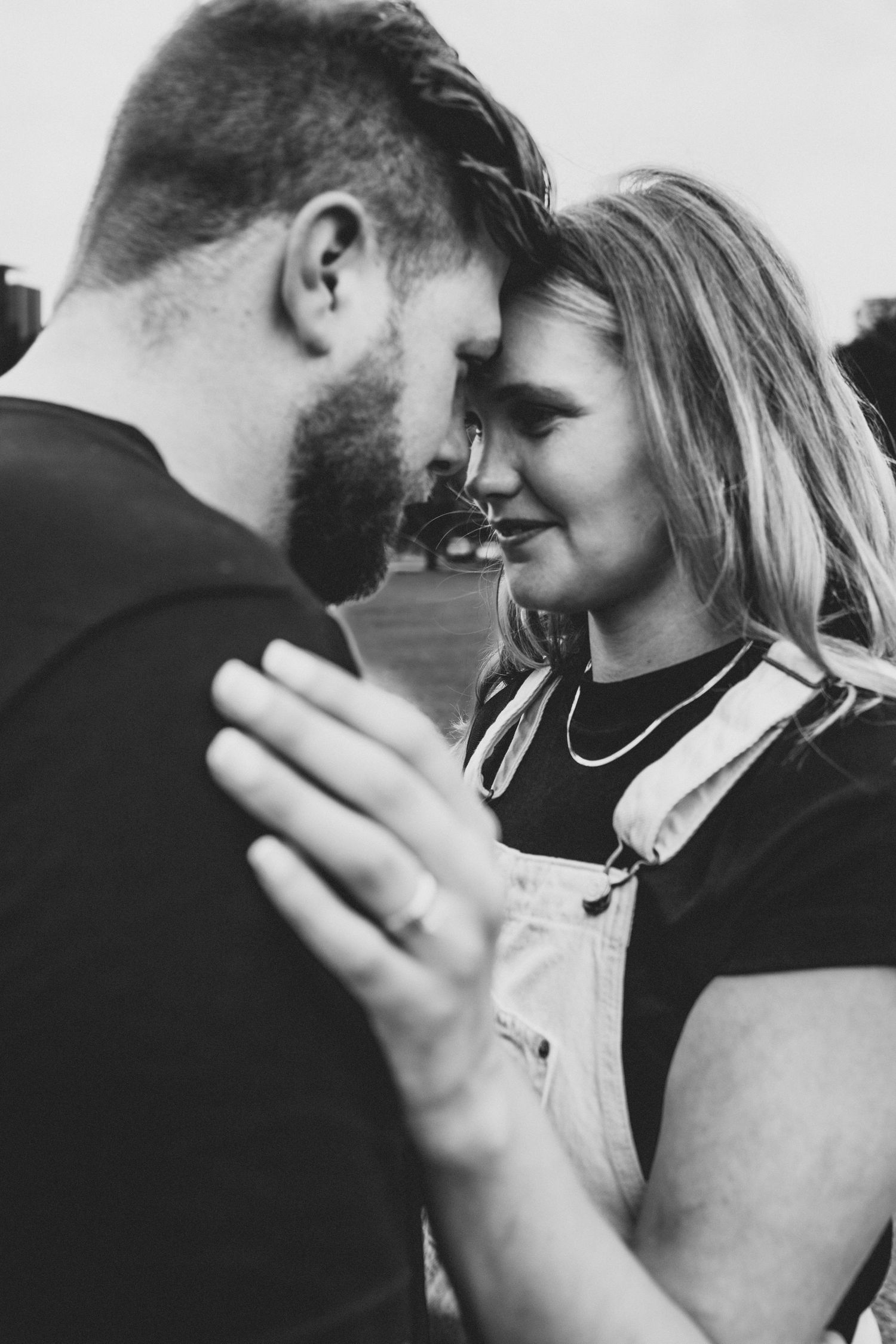 Black and white photo of a couple touching foreheads intimately, woman wearing overalls and showing engagement ring.