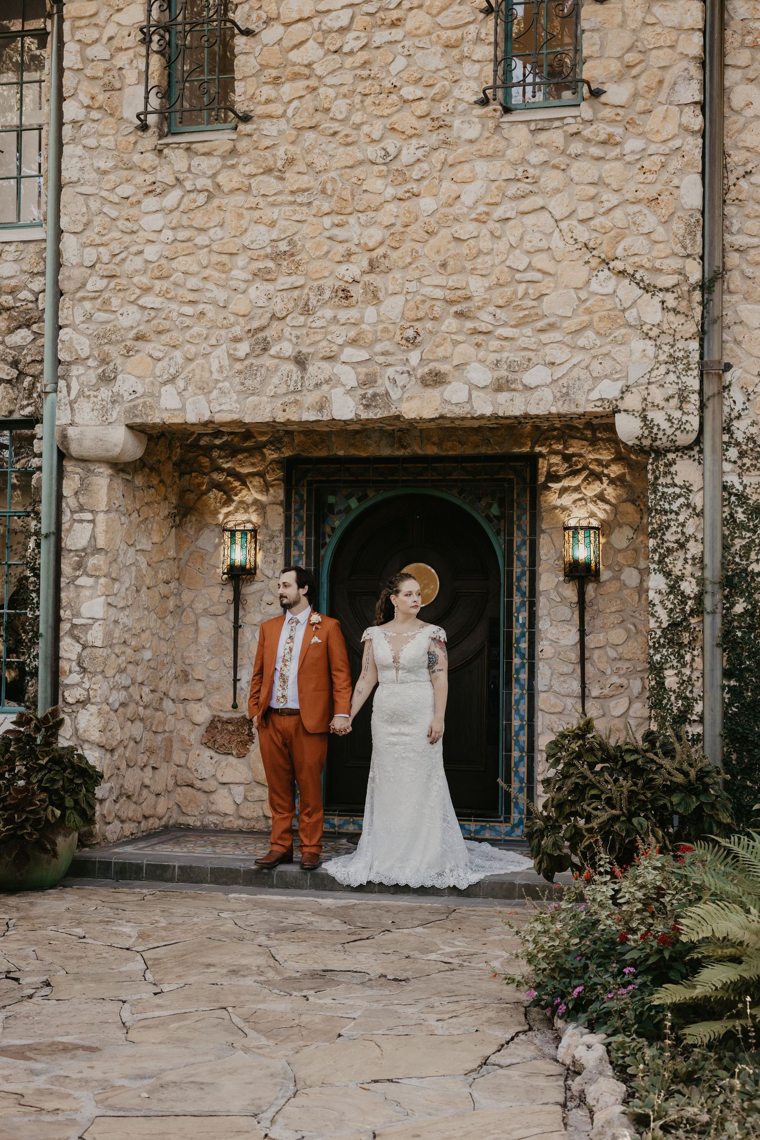 Bride and groom hold hands standing before an arched stone doorway at their elegant wedding venue.