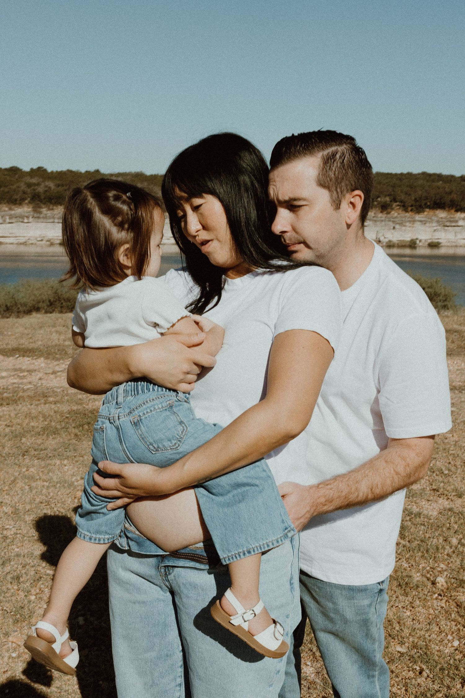 Family of three in white tops and jeans sharing a tender moment near a sunny lakeside.