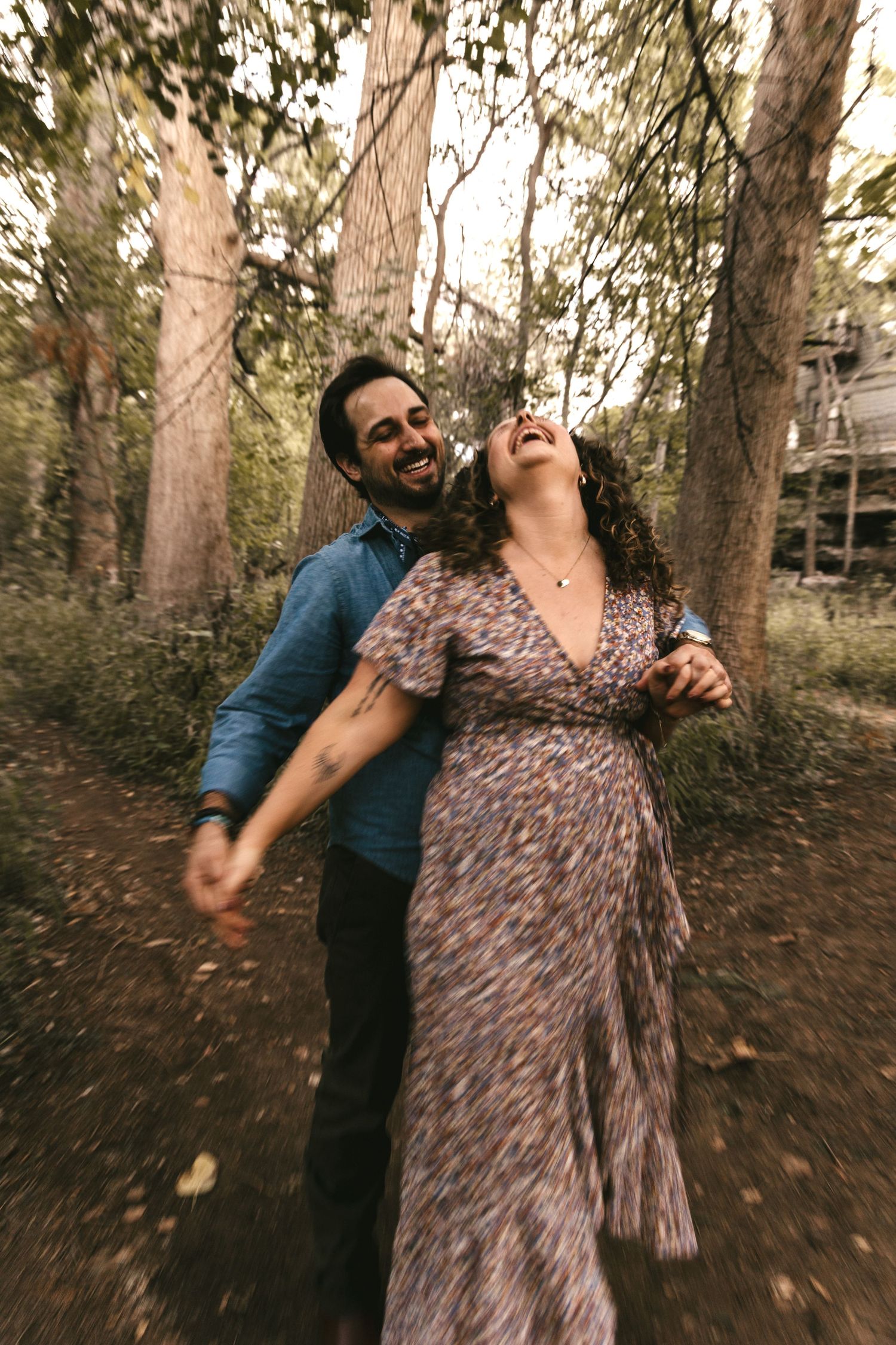 Joyful couple laughing and dancing together on a forest path surrounded by tall trees.