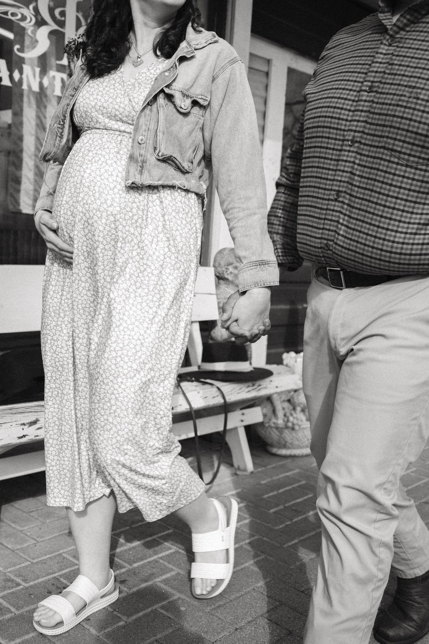 Black and white photo of a pregnant woman in a floral dress and denim jacket holding hands with a man outside a shop.